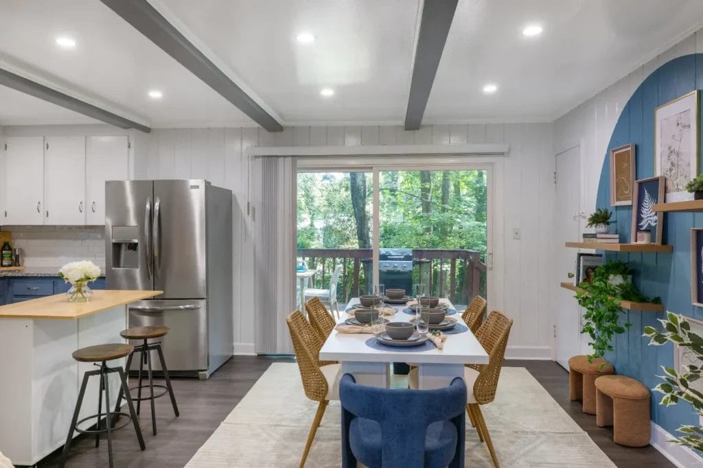 Modern dining area with a set table for six, woven chairs, and a blue accent wall with shelves. The space has a kitchen with stainless steel appliances and large glass doors revealing a green outdoor view.