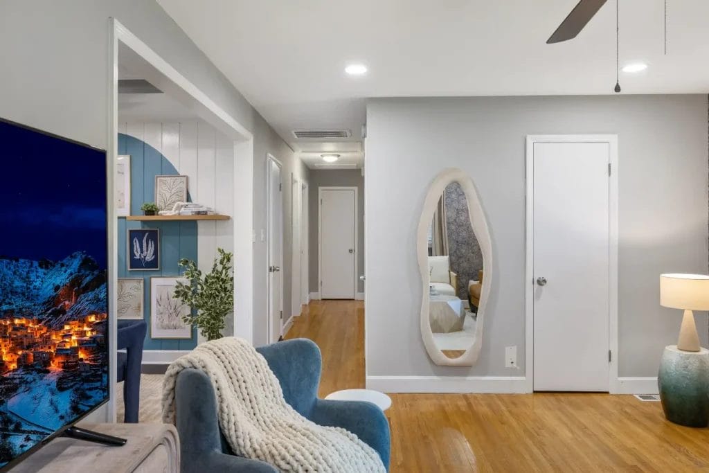 A cozy living room with a blue armchair, a textured blanket, a TV showing a snowy village, wooden floors, a hallway, a wavy wall mirror, a lamp, and decorated shelves with books and plants.