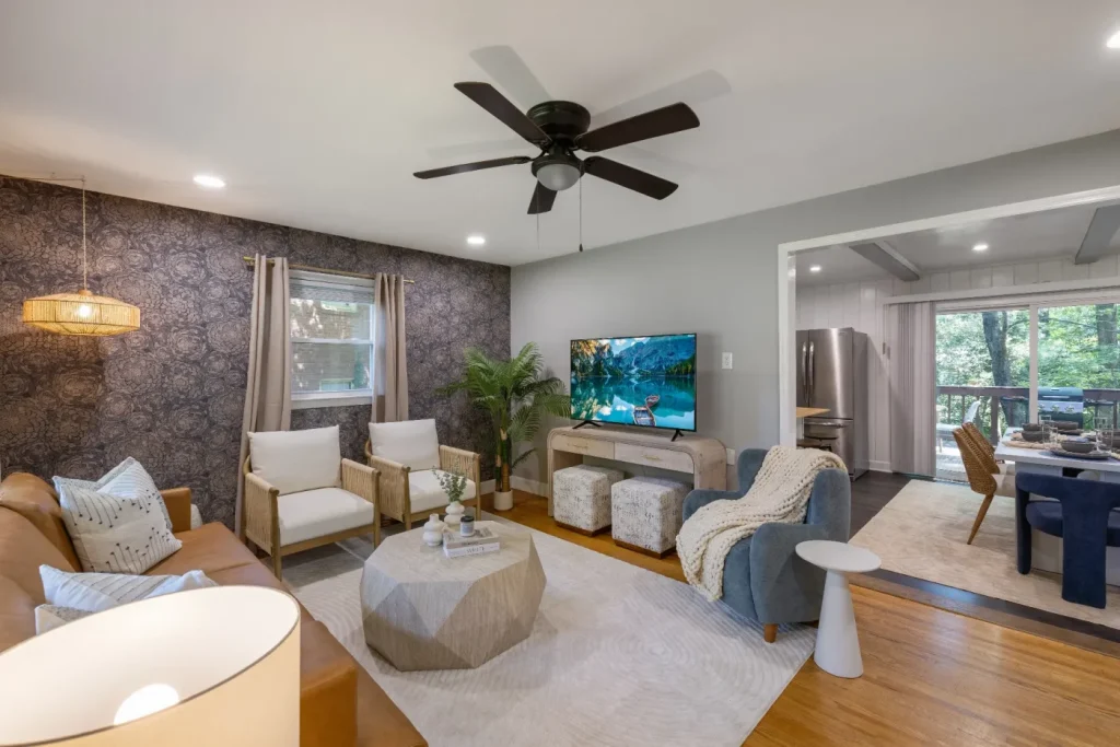 Modern living room with a ceiling fan, brown leather sofa, two white chairs, TV on the wall, poufs, geometric coffee table, and a dining area and kitchen visible in the background; natural light from windows.