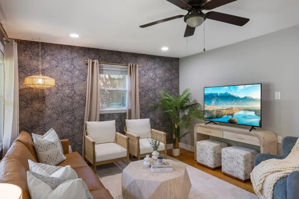 A cozy living room with a brown leather sofa, two white armchairs, a round wooden coffee table, a TV on a console, potted plant, patterned accent wall, ceiling fan, and large window with beige curtains.