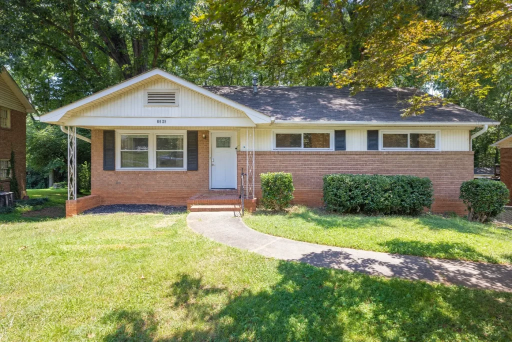 Single-story brick house with white trim, a covered front porch, and a curved walkway leading to the front steps. The yard is grassy with bushes and large trees providing shade in the background.