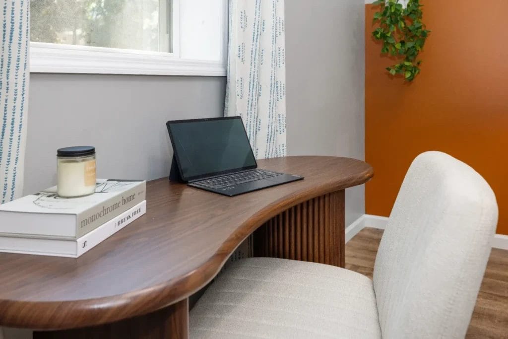 A modern home office desk with a closed laptop, a candle, and two stacked books beside a cushioned chair. There’s a window, white and blue curtains, orange accent wall, and a potted plant in the background.