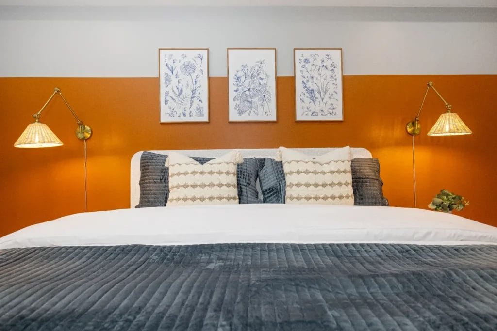 A neatly made bed with patterned pillows, a gray blanket, and two wall-mounted lamps on either side. The wall behind the bed is orange with three framed floral prints hanging above the headboard.