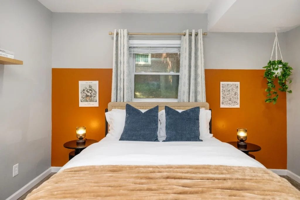A cozy bedroom with a bed featuring white linens, blue accent pillows, and a tan blanket. Two bedside tables with lamps, art on the orange accent wall, curtains, a window, and a hanging plant add warmth to the space.