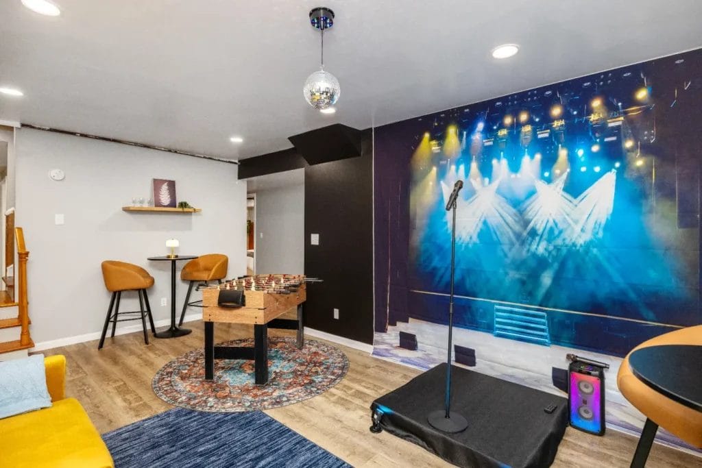 A modern rec room with a foosball table, small round tables, yellow couch, disco ball, and a stage setup with a microphone and speaker. The back wall features a mural of a concert stage with colorful spotlights.