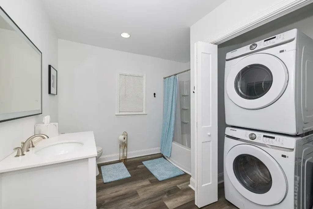 Modern bathroom with white walls and wood flooring, featuring a vanity with sink, toilet, shower with blue curtain, blue rugs, and a stacked washer and dryer next to a closet with an open white door.