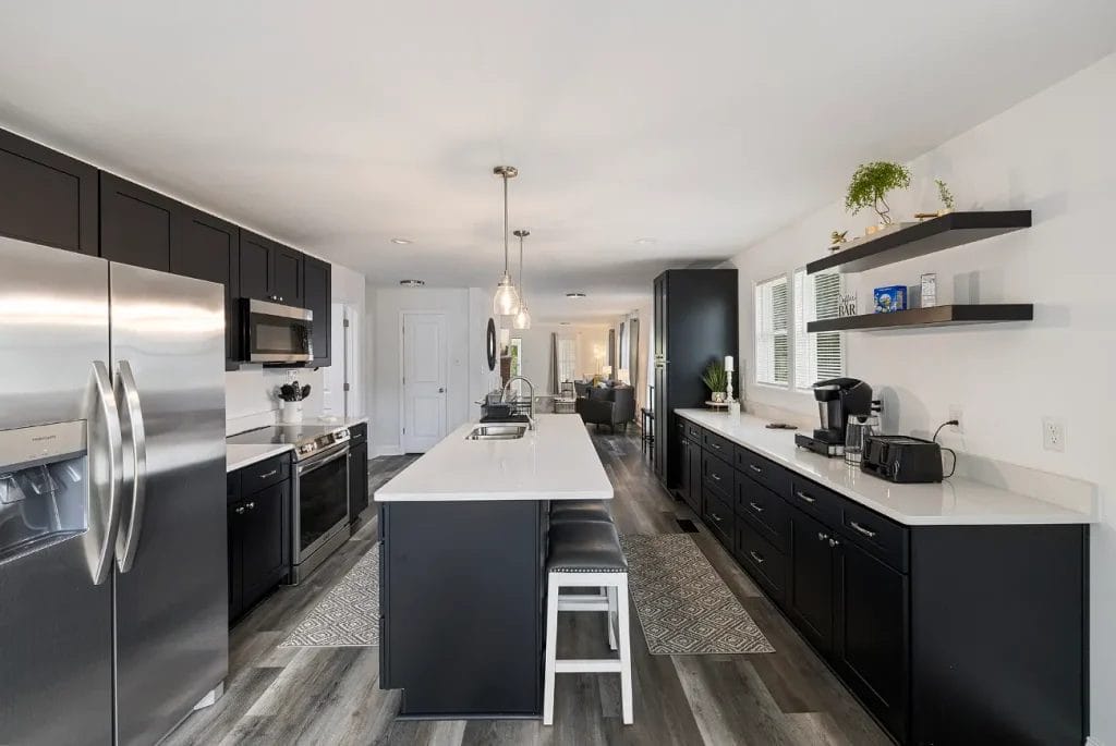 Modern kitchen with black cabinets, a large white island with barstools, stainless steel appliances, open shelves with decor, and wood-look flooring. Bright space with ample lighting and a view into the living area.