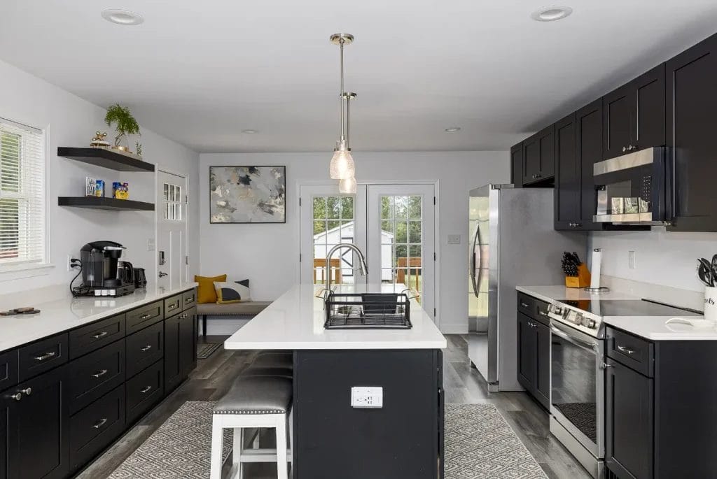 Modern kitchen with black cabinets, a white island and countertops, stainless steel appliances, pendant lighting, and a cozy breakfast nook by French doors leading outside. A coffee station and decorative accents complete the space.