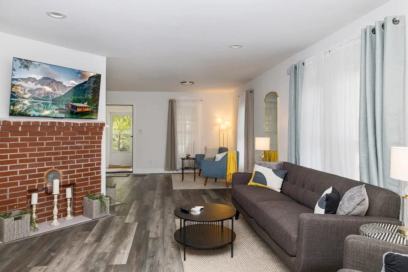 A modern living room with a gray sofa, round black coffee table, and a brick fireplace. A TV is mounted above the fireplace. Light gray curtains cover the windows, and a blue armchair with a floor lamp sits in the corner.