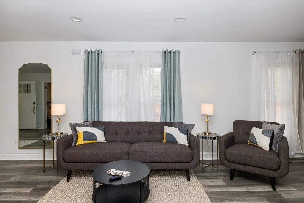 A modern living room with a dark gray sofa, matching armchair, round black coffee table, side tables with lamps, and light curtains over two windows. The decor features gray, white, and yellow accent pillows.