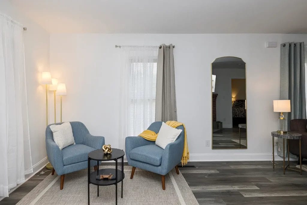 Two blue armchairs with white pillows and a yellow blanket sit around a small black round table in a modern living room with floor lamp, large mirror, and neutral decor. A gray rug covers part of the wood floor.