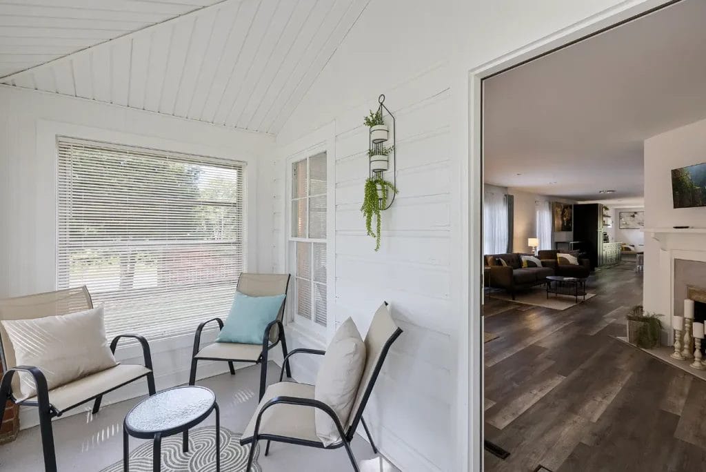 A bright sunroom with three cushioned chairs around a small glass table, large windows with blinds, potted plants on the wall, and an open doorway leading to a modern living room with wood flooring.