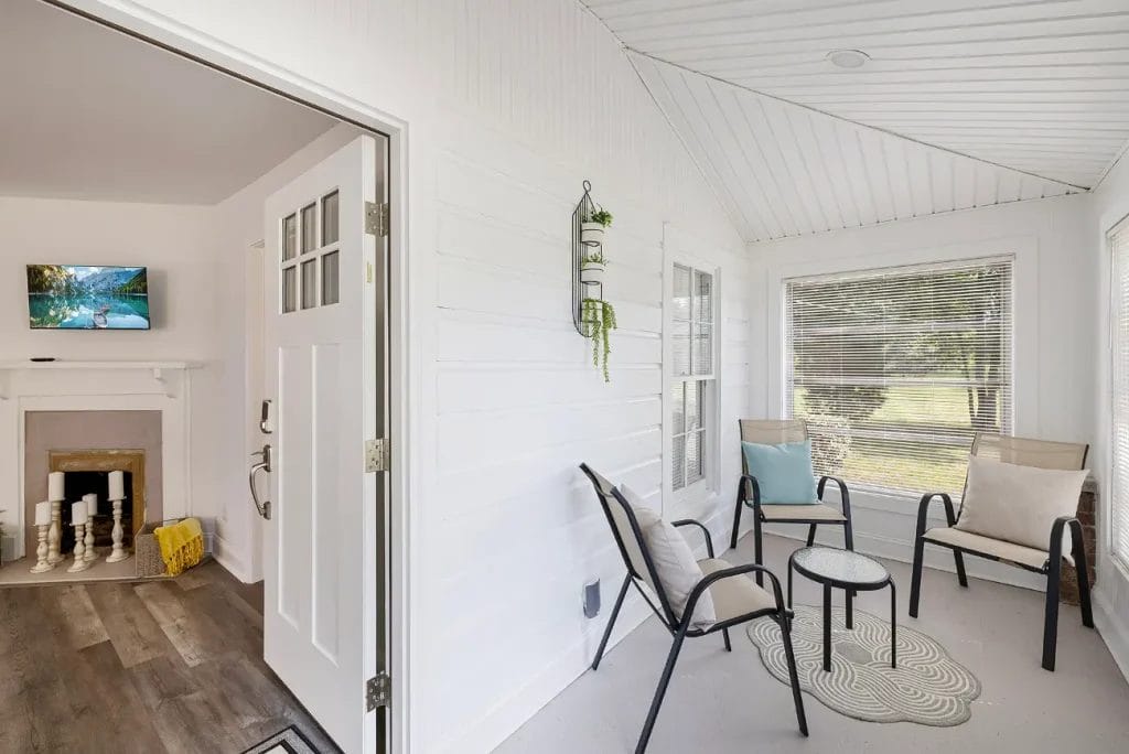 A bright sunroom with white walls, three chairs, a small round table, and large windows. An open door leads to a living room with a fireplace, television, and wooden floors.
