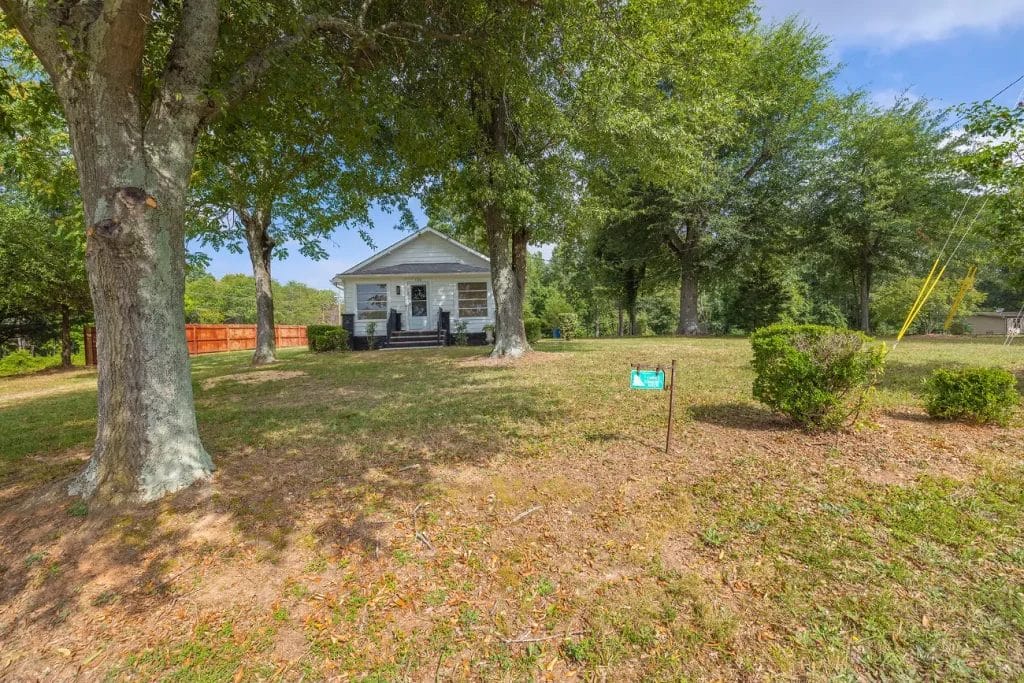 A small white house with a front porch sits shaded by large trees on a spacious grassy lawn. A wooden fence and bushes are visible, with a sign in the yard near the front. The sky is partly cloudy.