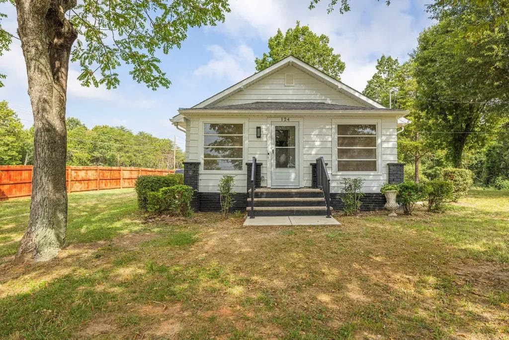A small white house with a covered front porch, black steps, and trim. The yard has grass, bushes, and a tree, with a red wooden fence on the left and dense green trees in the background.