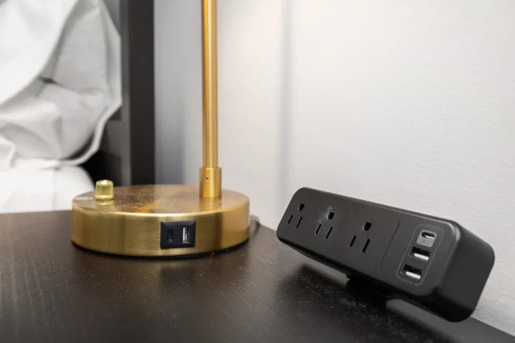 A gold lamp with a USB charging port on its base sits on a black nightstand next to a black power strip with three outlets and two USB ports, against a white wall.