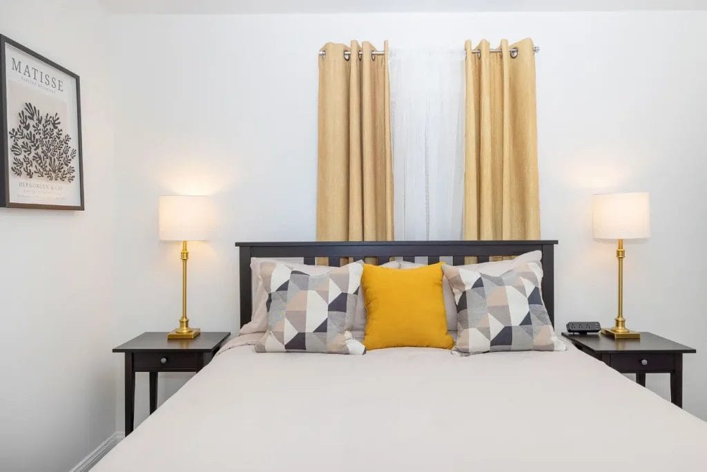 A neatly made bed with geometric-patterned and yellow pillows sits between two black nightstands with gold lamps. Yellow curtains and a framed Matisse print decorate the white wall behind the bed.