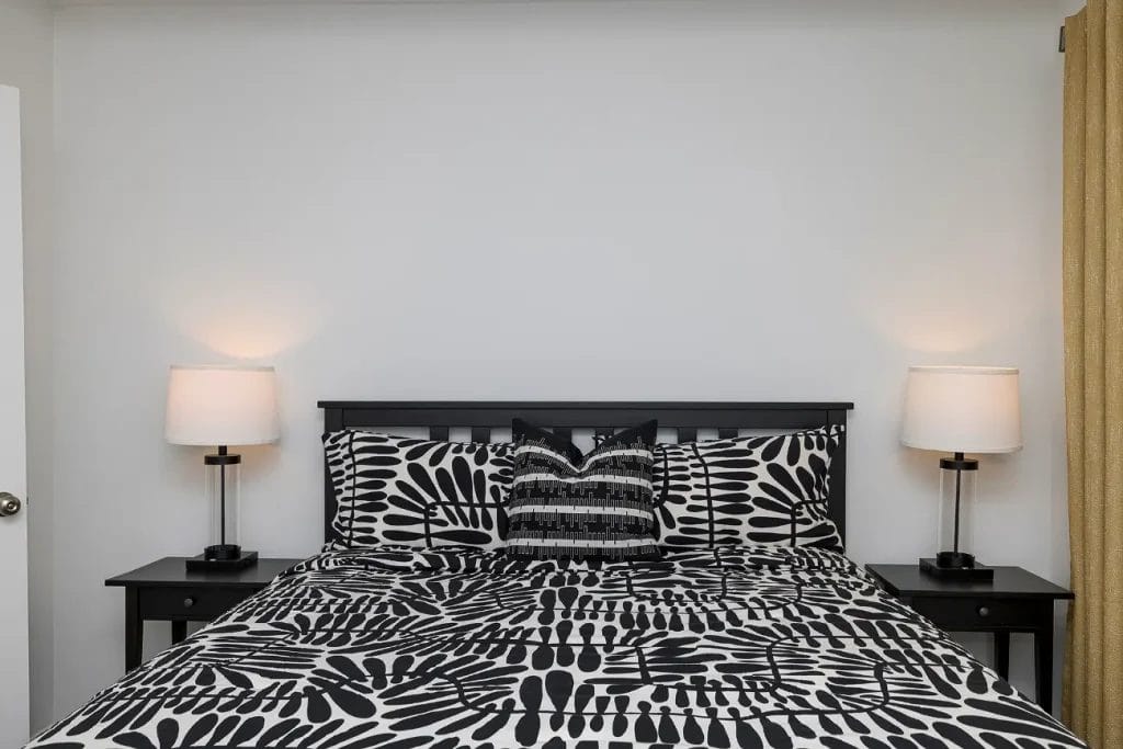 A neatly made bed with black and white patterned bedding is centered between two black nightstands, each with a lamp. The wall behind the bed is plain and white, and beige curtains hang on the right side.