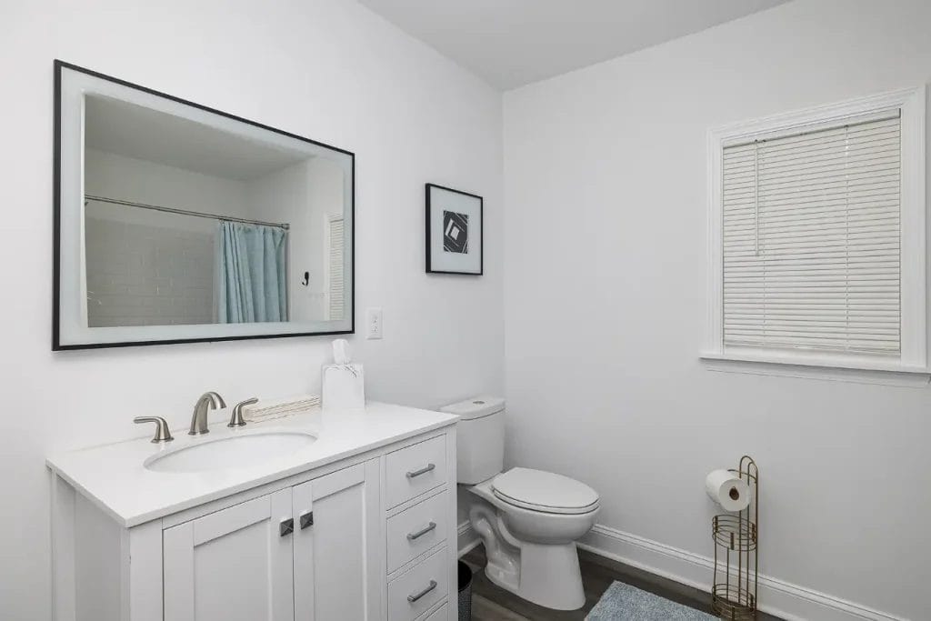 A modern, white bathroom with a vanity and sink, large mirror, toilet, toilet paper stand, window with blinds, framed art, and a shower with a blue curtain. The space is clean and minimalistic.