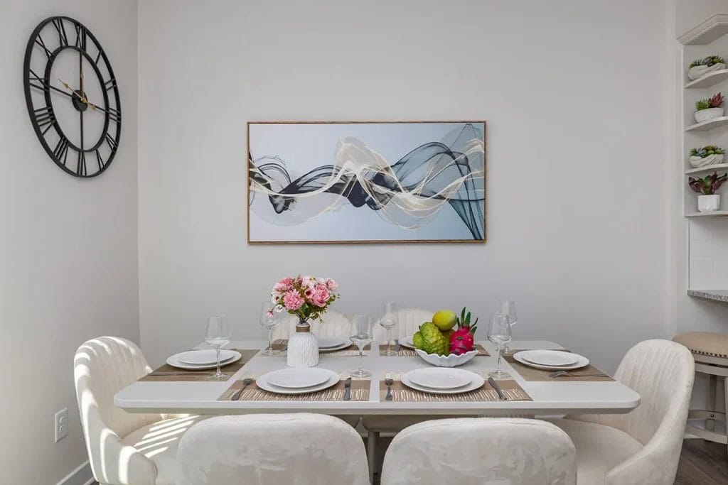A dining table set for four with white chairs, plates, glasses, and a flower centerpiece. On the wall are a modern clock and abstract artwork. A bowl of fruit and a dragon fruit are on the table. Shelves with dishes are nearby.