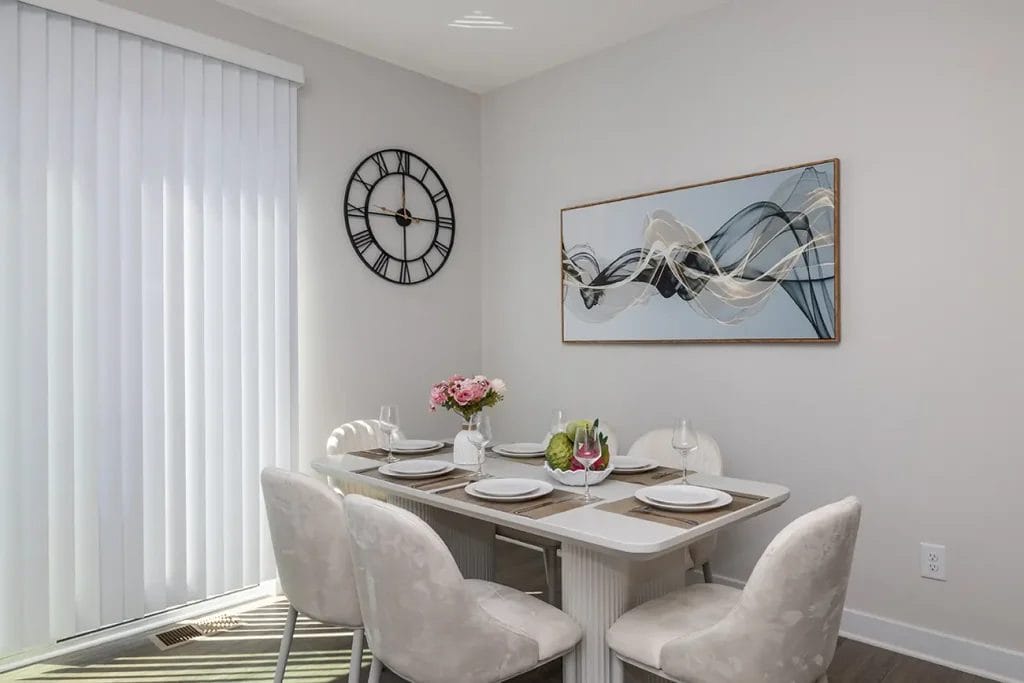 A modern dining area with a white table set for four, cream upholstered chairs, a vase of pink flowers, abstract wall art, a large black clock, and vertical blinds letting in natural light.