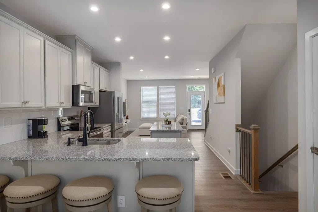 Modern kitchen with gray cabinets and granite countertops, island with four cushioned stools, stainless steel appliances, and an open view to a bright living and dining area with large windows and wood flooring.