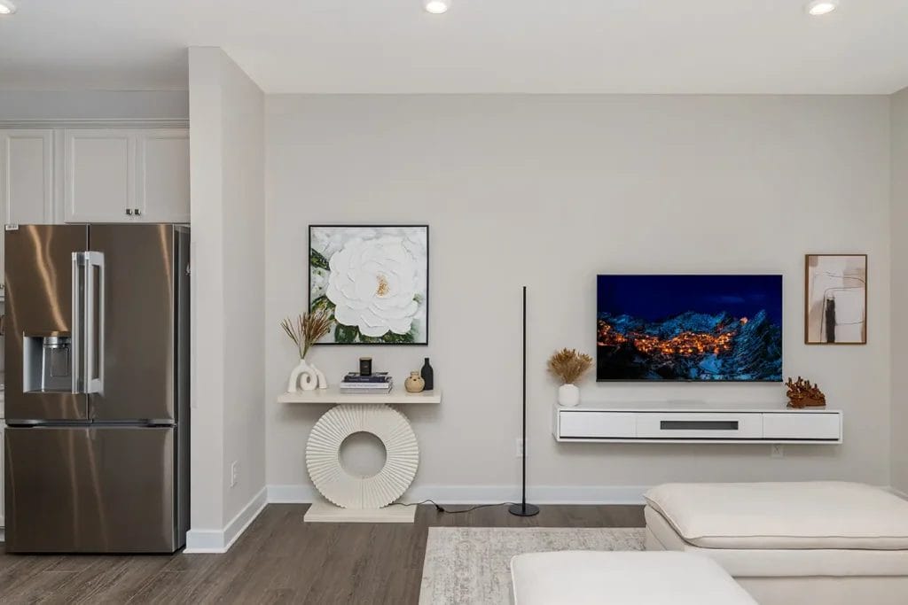 Modern living room with a mounted TV displaying a night mountain scene, a white console table with decor, wall art, a tall black floor lamp, and part of a stainless steel refrigerator visible on the left.