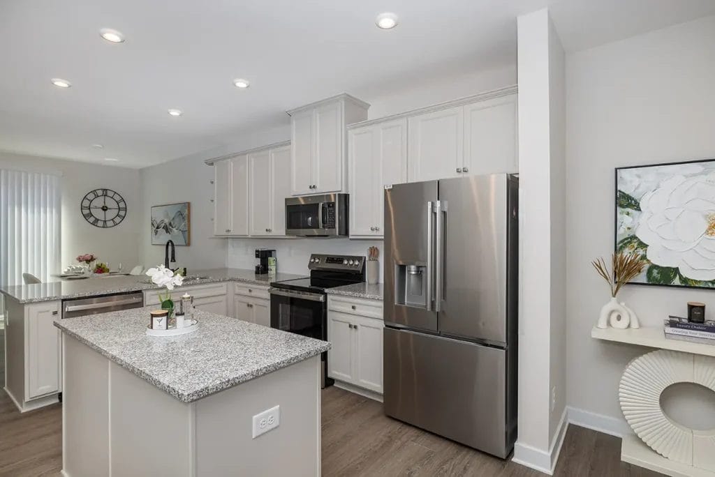 Modern kitchen with light gray cabinets, stainless steel appliances, granite countertops, a center island, wood flooring, and minimalist decor including a flower painting and a wall clock in the background.
