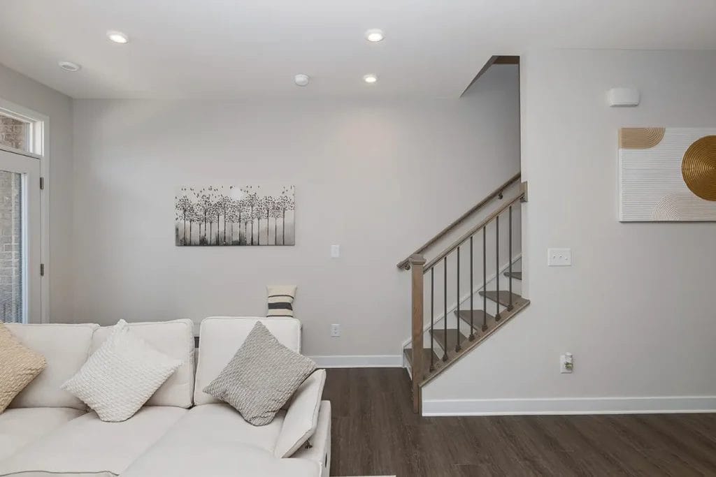 A modern living room with a white sofa, neutral-toned pillows, wooden flooring, a staircase with metal railings, and minimal wall art on light gray walls.