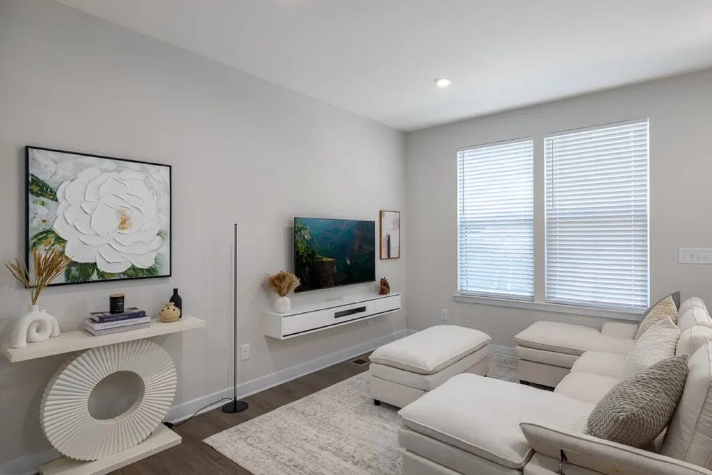 A modern living room with a white sectional sofa, ottomans, a wall-mounted TV, and large windows with blinds. Minimalist decor includes a console table with a flower painting and decorative items.