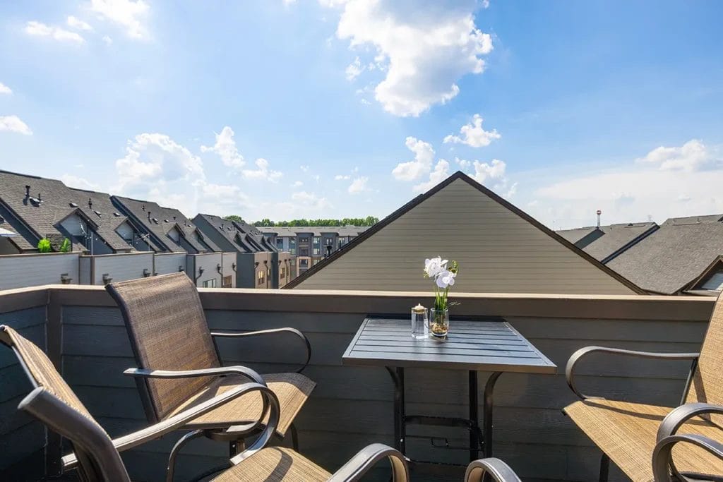 A small balcony with two chairs and a table holding a vase of white flowers overlooks rows of modern townhouses under a bright blue sky with scattered clouds.