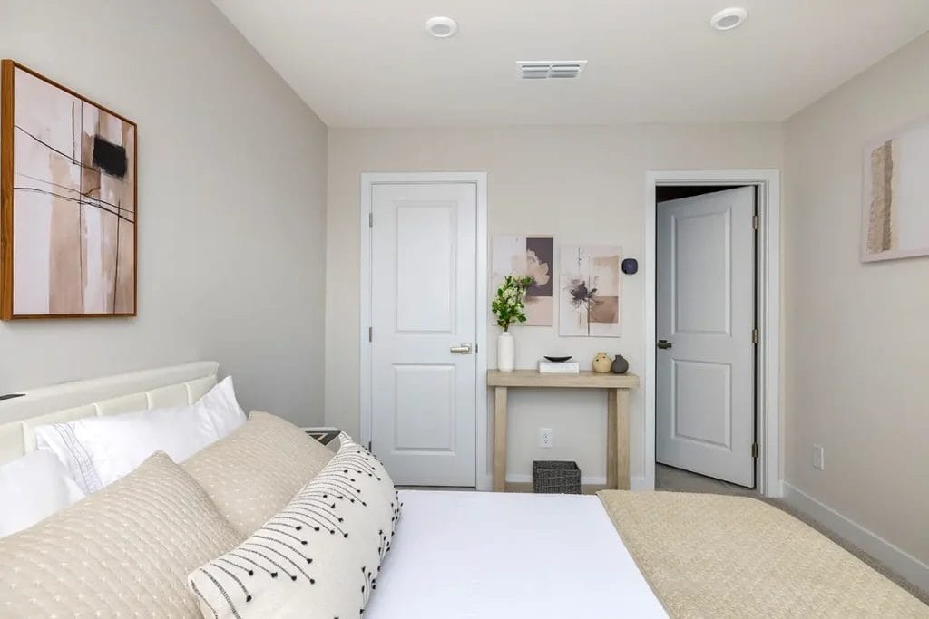 A modern bedroom with a neatly made bed, beige and white bedding, abstract art on cream walls, a wooden console table with decor, and two white doors, one open and one closed.