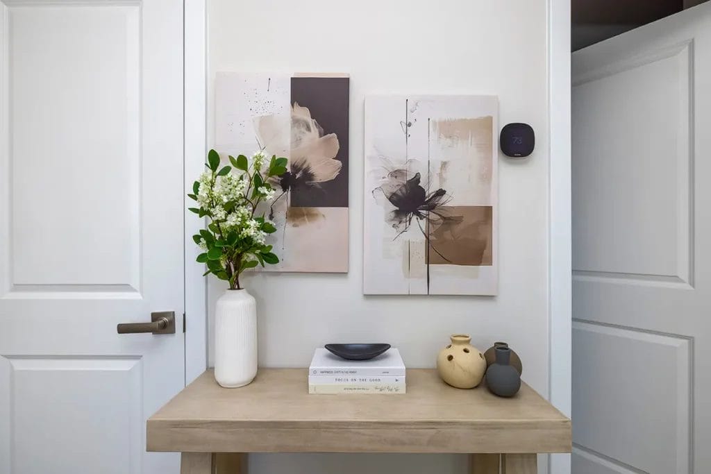 A minimalist entryway with a light wooden console table, two abstract art pieces on the wall, a white vase with green foliage, three decorative vases, a stack of books, and a smart thermostat on the wall.