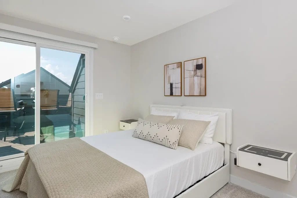 A modern bedroom with a neatly made bed featuring beige bedding, two abstract art pieces above the headboard, and sliding glass doors leading to a balcony with patio chairs and a table.