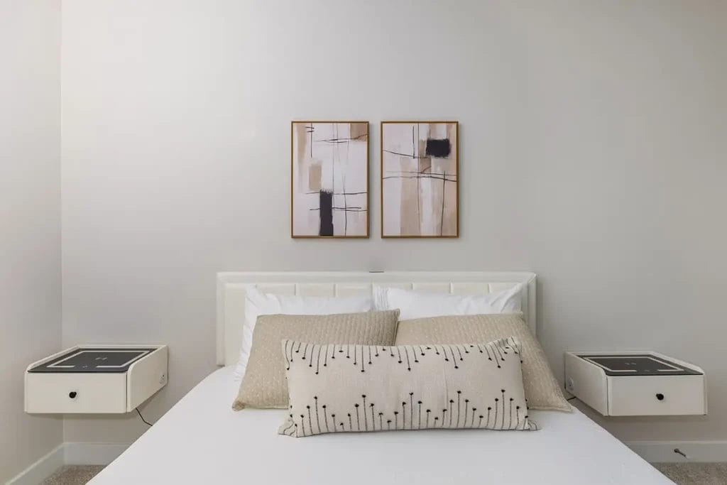 A minimalistic bedroom with a white bed, two beige pillows, a patterned bolster, two floating nightstands, and two abstract paintings hanging above the headboard on a light-colored wall.