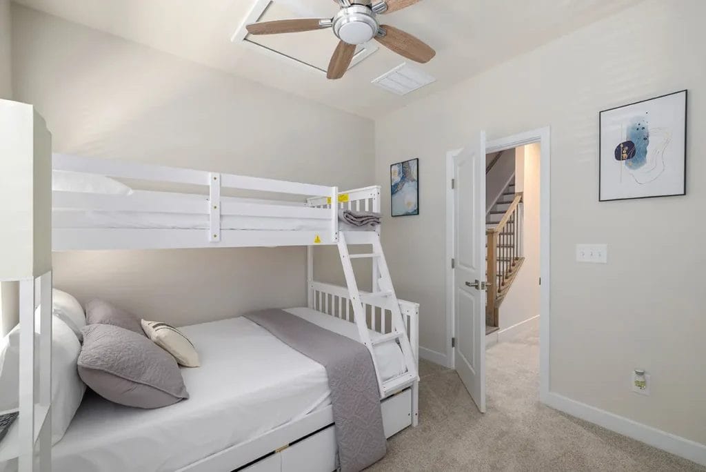 A bright bedroom with a white bunk bed, light bedding, a ceiling fan, beige walls, carpeted floor, and two framed artworks. An open door reveals a staircase and hallway outside the room.