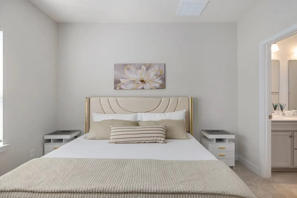 A modern, neutral-toned bedroom with a beige upholstered bed, striped and solid pillows, two white nightstands, a floral wall art above the headboard, and an open doorway leading to a well-lit bathroom.