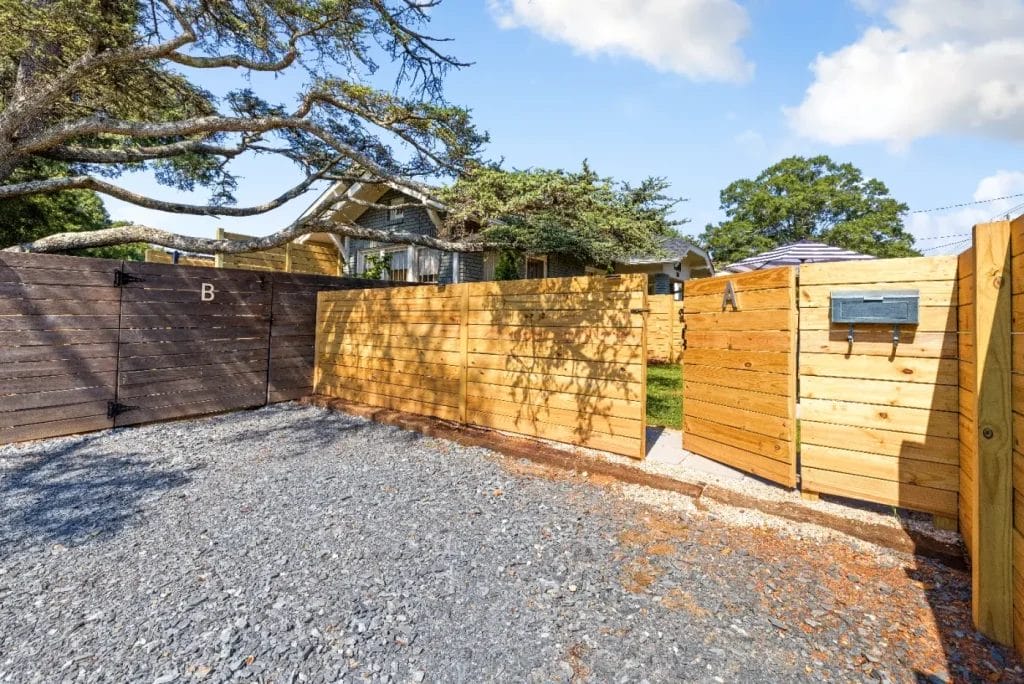 Wooden fence with a gate and mailbox encloses a gravel driveway. Tree branches hang over the fence, and a house with a porch is visible in the background under a partly cloudy sky.