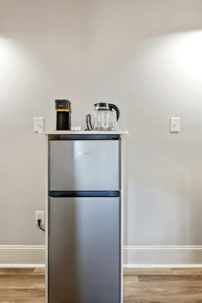 A small silver mini fridge stands against a light gray wall with a white countertop holding a black coffee maker, a glass coffee pot, stacked glass cups, and a spoon. Two empty wall switches are visible on either side.