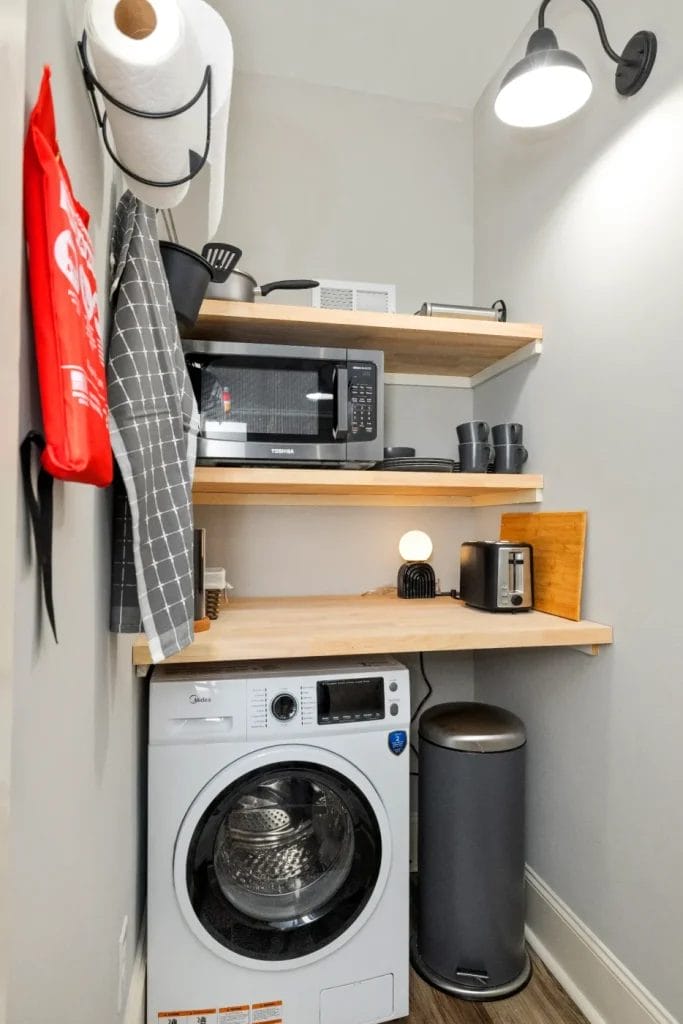 A compact laundry room with a washing machine, trash can, wooden shelves holding a microwave, dishes, toaster, lamp, utensils, paper towels, apron, and a checked towel hanging on the wall under a black wall lamp.