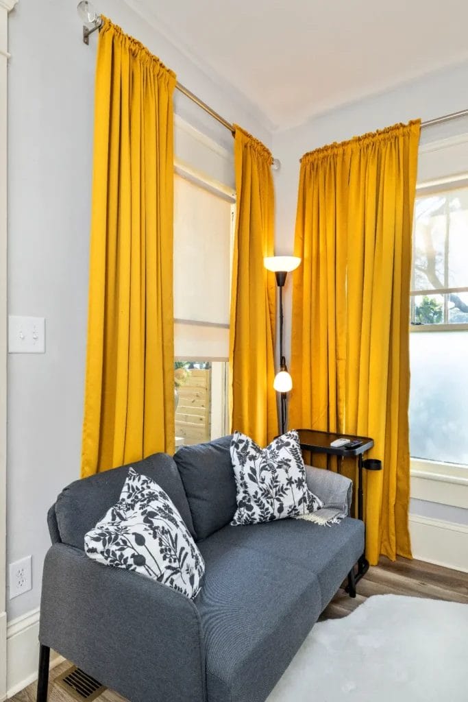 A small gray sofa with black and white patterned pillows sits in a corner by two windows with bright yellow curtains. A black floor lamp and a small side table are next to the sofa. The room has light-colored walls and flooring.