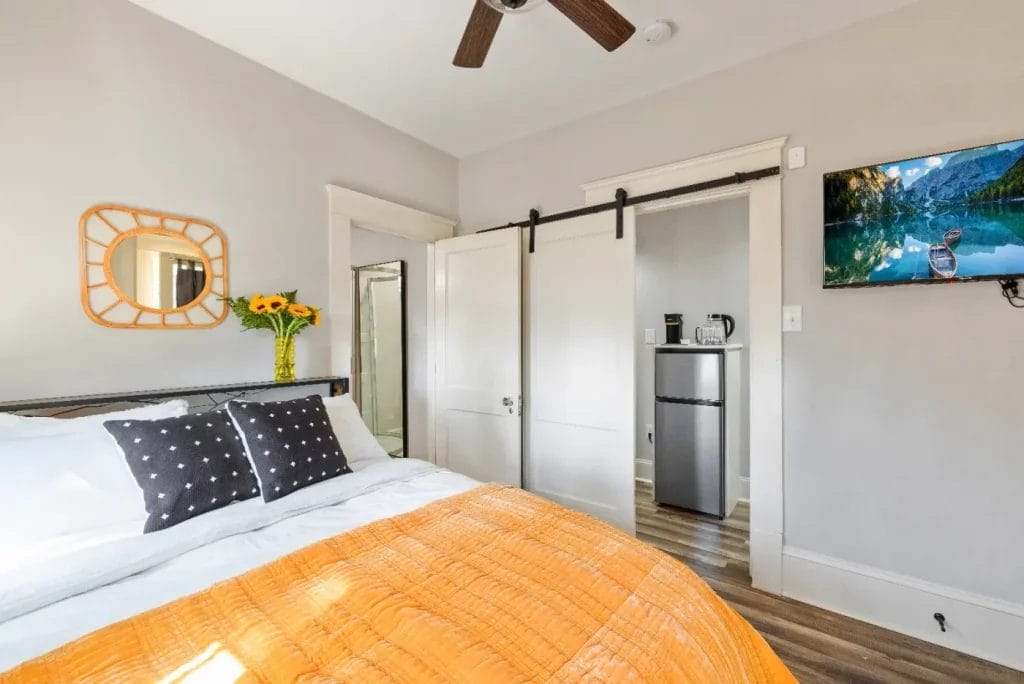 Bright bedroom with a bed covered in an orange blanket, black-and-white pillows, a vase of sunflowers, a wall mirror, sliding barn doors, a ceiling fan, a mini fridge, and a wall-mounted TV displaying a nature scene.