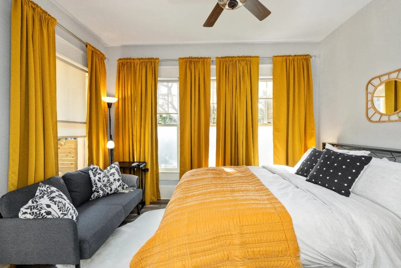 A bright bedroom with three large windows covered by gold curtains, a bed with a gold quilt and black-and-white pillows, a gray sofa with patterned cushions, a floor lamp, and a ceiling fan.