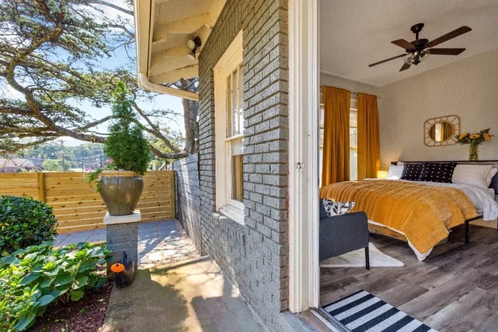 A cozy bedroom with mustard yellow bedding opens onto a small patio with foliage, a tall planter, and a wooden fence under a sunny sky. The room features a ceiling fan, gray brick exterior, and large windows with yellow curtains.