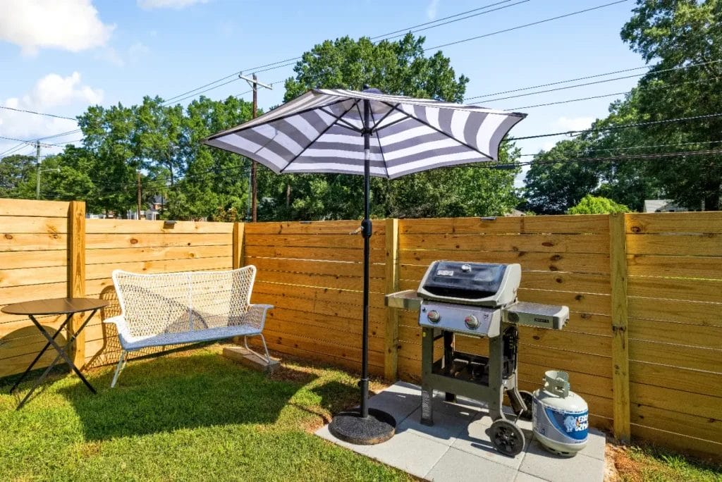 A backyard patio with a wooden fence, a white woven bench, a small round table, a black-and-white striped umbrella, and a gas grill with a propane tank on a concrete pad. Trees are visible in the background.