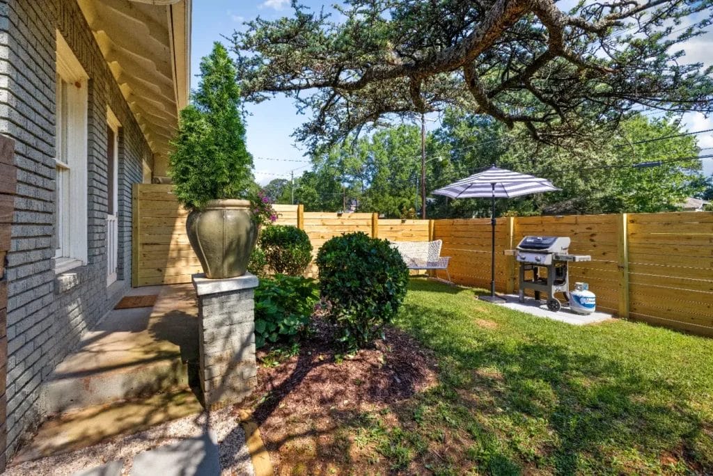 A small fenced backyard with green grass, neatly trimmed bushes, a grill under a striped umbrella, a bench, and a large potted plant near the house. Sunlight filters through a large tree overhead.