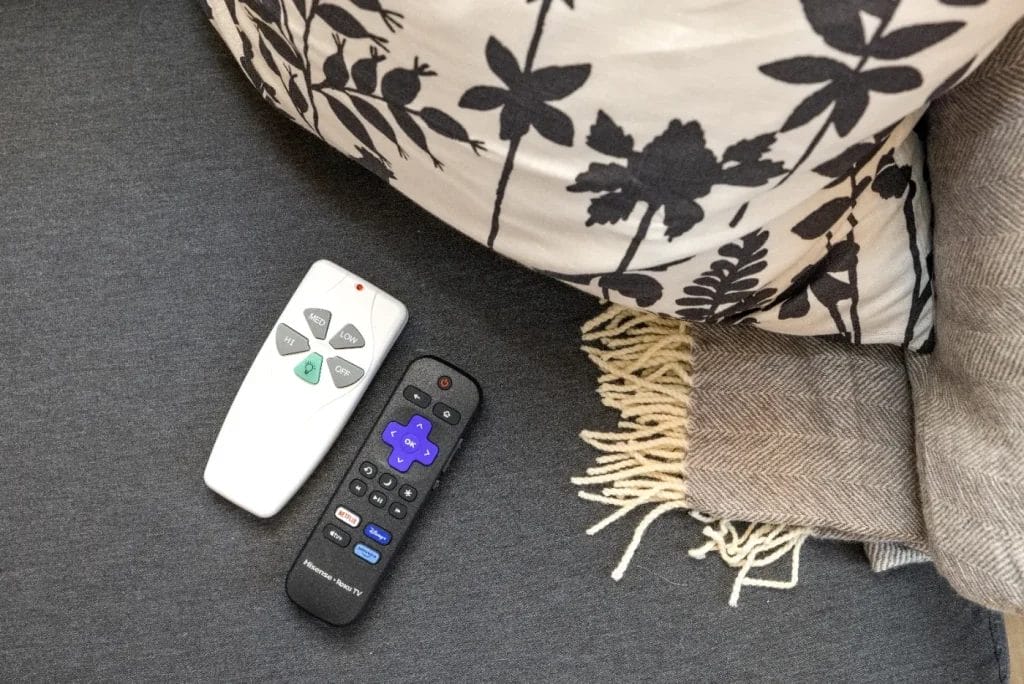 Two remote controls, one white and one black, are placed on a gray surface next to a pillow with a black floral pattern and a beige fringed blanket.