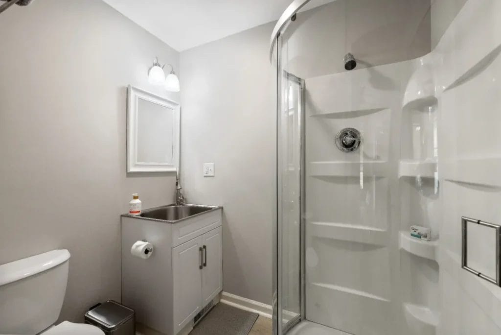 A modern bathroom with a glass-enclosed corner shower, a white vanity with a sink, a mirror above, and a toilet next to a small trash can under bright lighting.