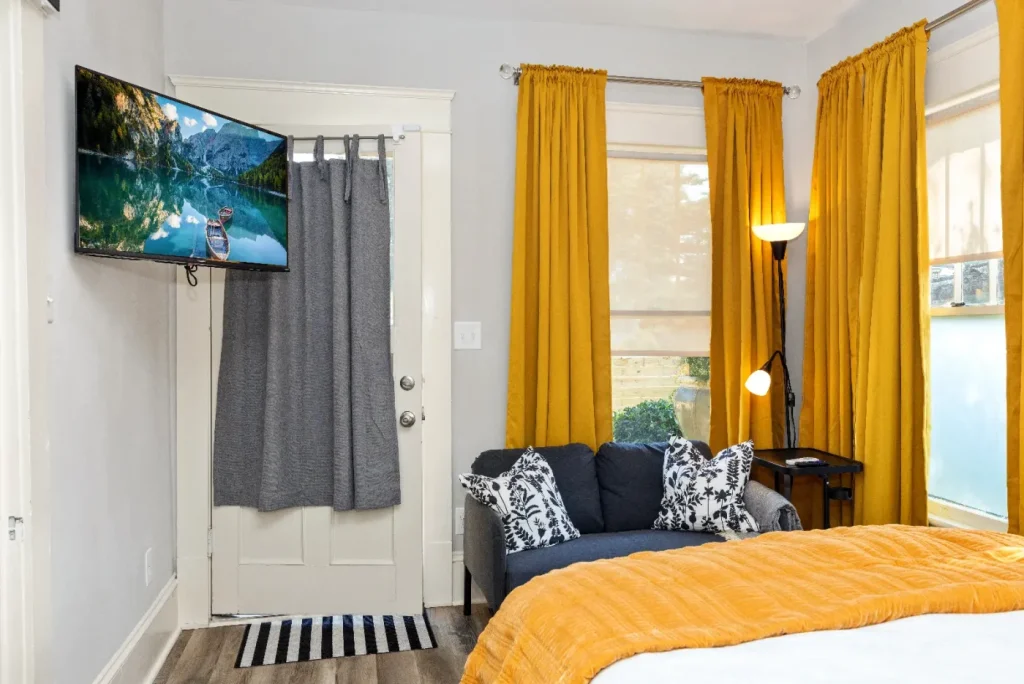 A cozy bedroom with mustard yellow curtains, a gray loveseat with patterned pillows, a floor lamp, a wall-mounted TV, and a bed with a mustard yellow comforter near a door with a gray curtain.