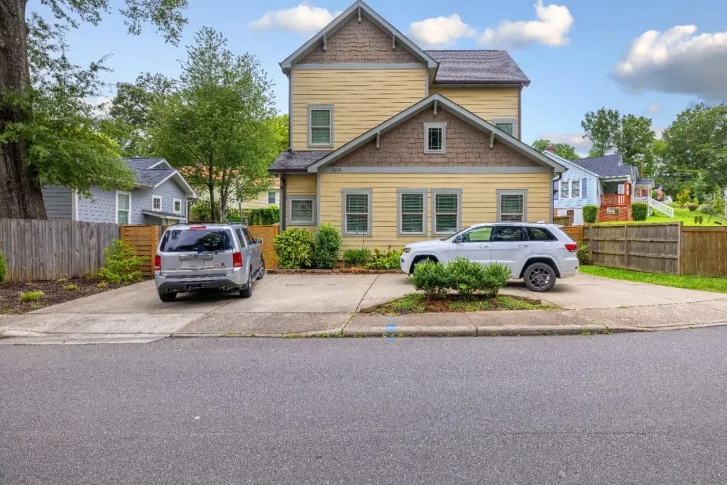 A two-story beige house with brown trim and green shutters stands behind two parked vehicles on a driveway, surrounded by a wooden fence, trees, and neighboring homes on a suburban street.